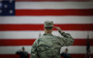 Amerykańscy lotnicy salutują podczas ceremonii przejęcia dowodzenia. Oryginalne zdjęcie z domeny publicznej Flickr. Na zdjęciu stoi żołnierz armii Stanów Zjednoczonych odwrócony do nas plecami. W tle widoczna jest flaga USA.